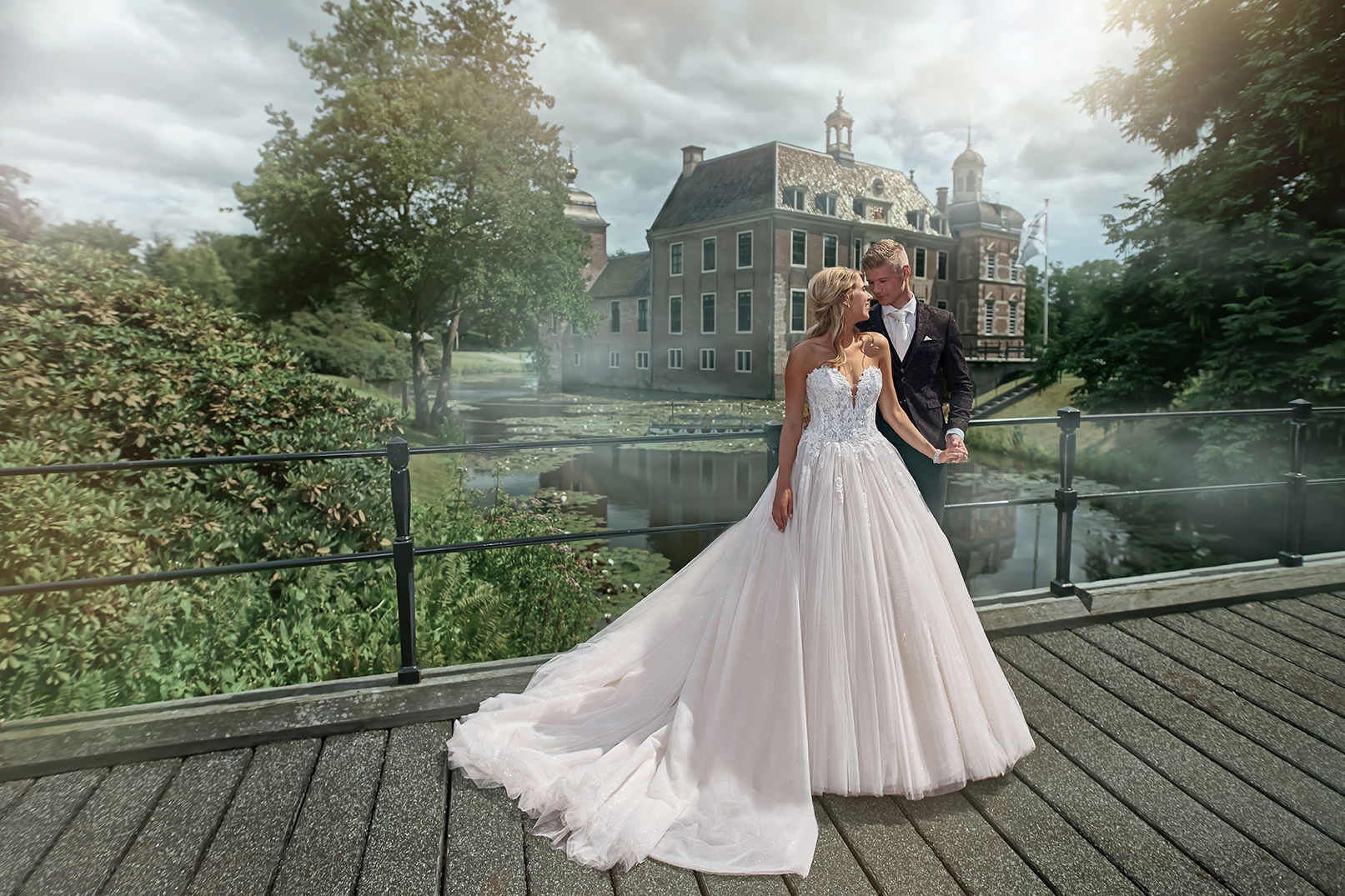 Braut und Bräutigam stehen vor einem prächtigen Herrenhaus auf einer Brücke. Die Braut trägt ein weites Kleid, und das Paar blickt sich verliebt an.