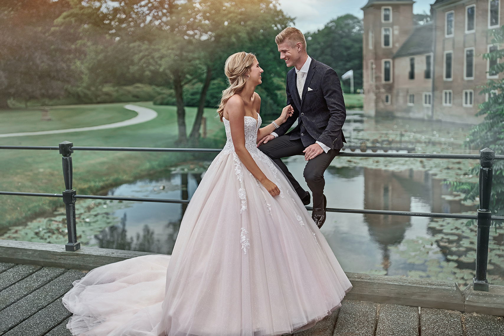 Braut und Bräutigam teilen einen liebevollen Moment auf einer Brücke. Die Braut trägt ein voluminöses Kleid, der Bräutigam sitzt am Geländer und lacht.