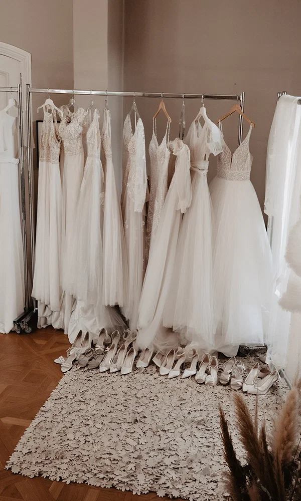 Eine Kleiderstange voller eleganter Brautkleider in einem hellen Raum. Auf dem Boden darunter sind zahlreiche Brautschuhe in verschiedenen Größen ordentlich aufgereiht.