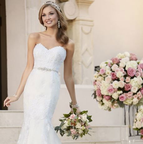 Strahlende Braut im trägerlosen Spitzenkleid von Stella York, mit Diadem und Brautstrauß, vor einer romantischen Szenerie aus rosafarbenen Blumenarrangements.