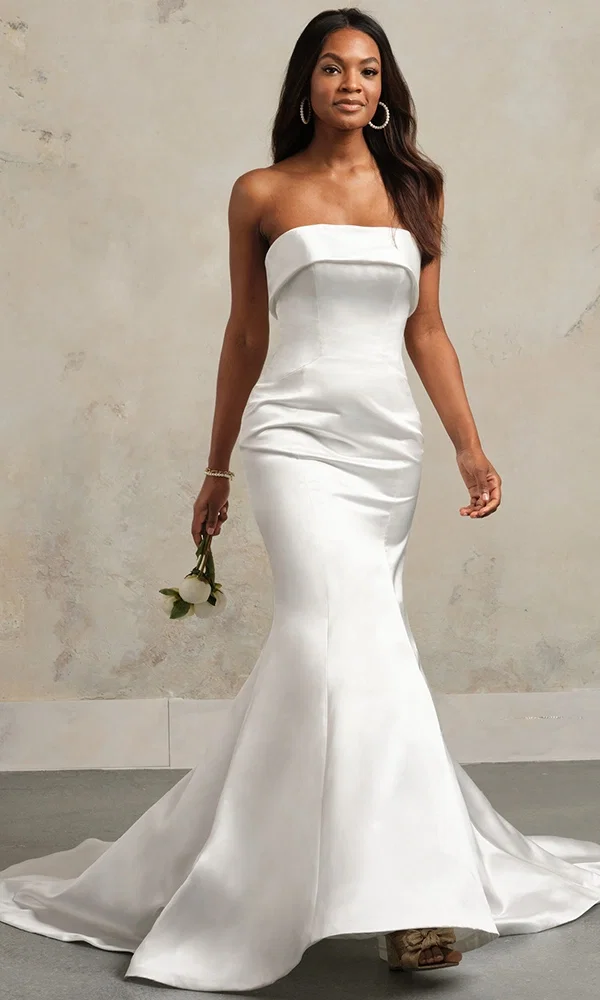 Braut in einem schlichten, eleganten Hochzeitskleid mit einer modernen, trägerlosen Silhouette, posiert in einem hell erleuchteten Raum mit klassischen Fliesen und großen Fenstern.
