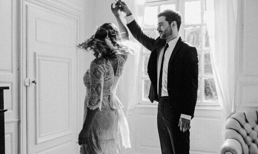 Romantischer Hochzeitstanz: Ein Bräutigam im Anzug führt seine Braut im Vintage-Spitzenkleid in einer eleganten Location mit hohen Fenstern.