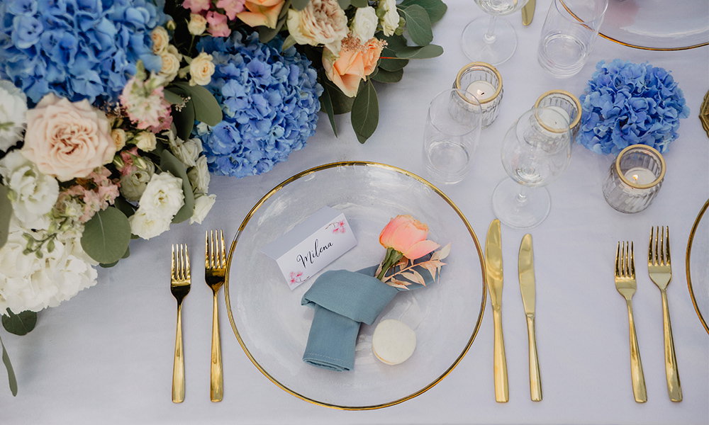 Festlich gedeckter Hochzeitstisch: Namenskarte auf Teller mit goldener Umrandung, dekoriert mit rosa Blüten, blauen Hortensien und sanftem Kerzenschein.