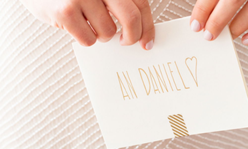 Eine Braut hält einen romantischen Briefumschlag mit handgeschriebener Widmung „An Daniel“ und einem goldenen Herz, auf hellem, strukturiertem Hintergrund.