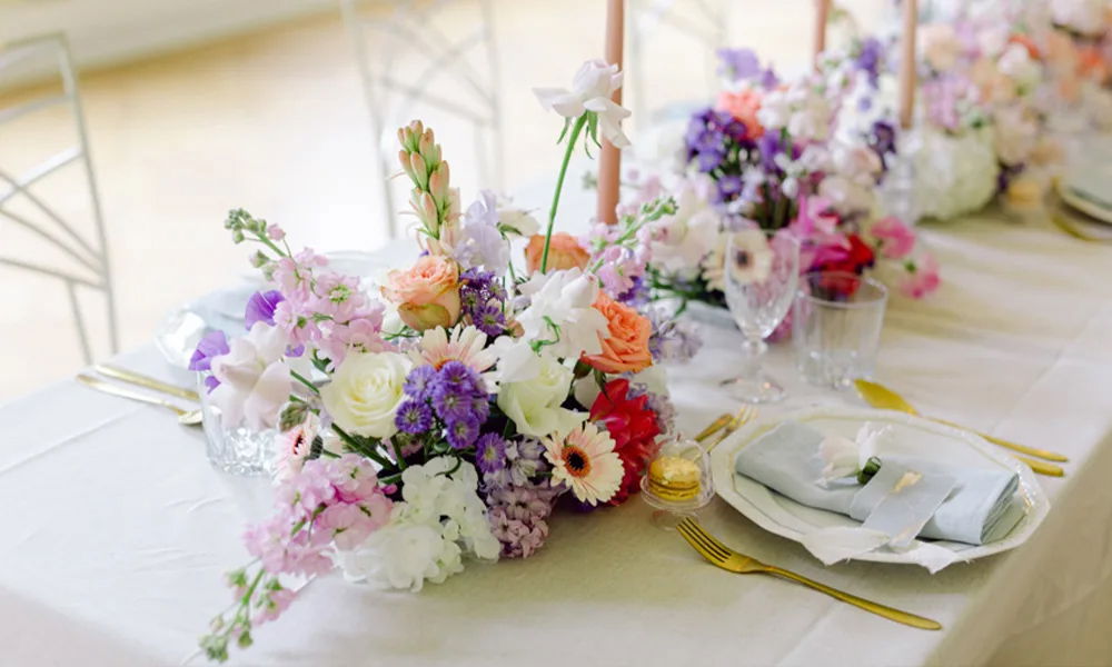 Hochzeitsgedeck mit romantischer Blumendeko in Rosa, Lila und Weiß, stilvollen Gläsern, goldfarbenem Besteck und kunstvoll gefalteten Servietten.