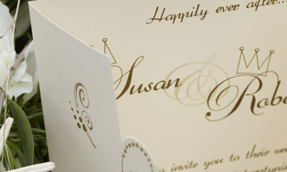 Klassische Hochzeitseinladung in Creme mit goldener Kalligrafie und den Namen „Susan & Robert“. Dekorative Kronen und zarte Muster schmücken das Design.