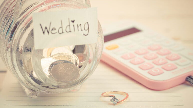 Ein mit Münzen gefülltes Glas mit 'Wedding'-Label, daneben ein rosafarbener Taschenrechner und ein Ring – Budgetplanung für die Hochzeit in Vorbereitung.