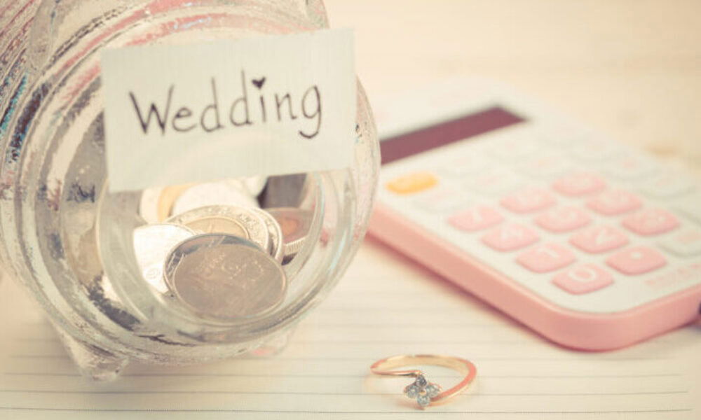 Ein mit Münzen gefülltes Glas mit 'Wedding'-Label, daneben ein rosafarbener Taschenrechner und ein Ring – Budgetplanung für die Hochzeit in Vorbereitung.
