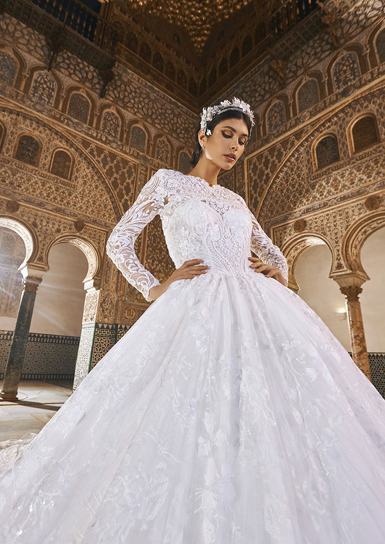 Prinzessinnenhaftes Brautkleid mit floralen Spitzen und langen Ärmeln, getragen in einem majestätischen, historischen Saal.