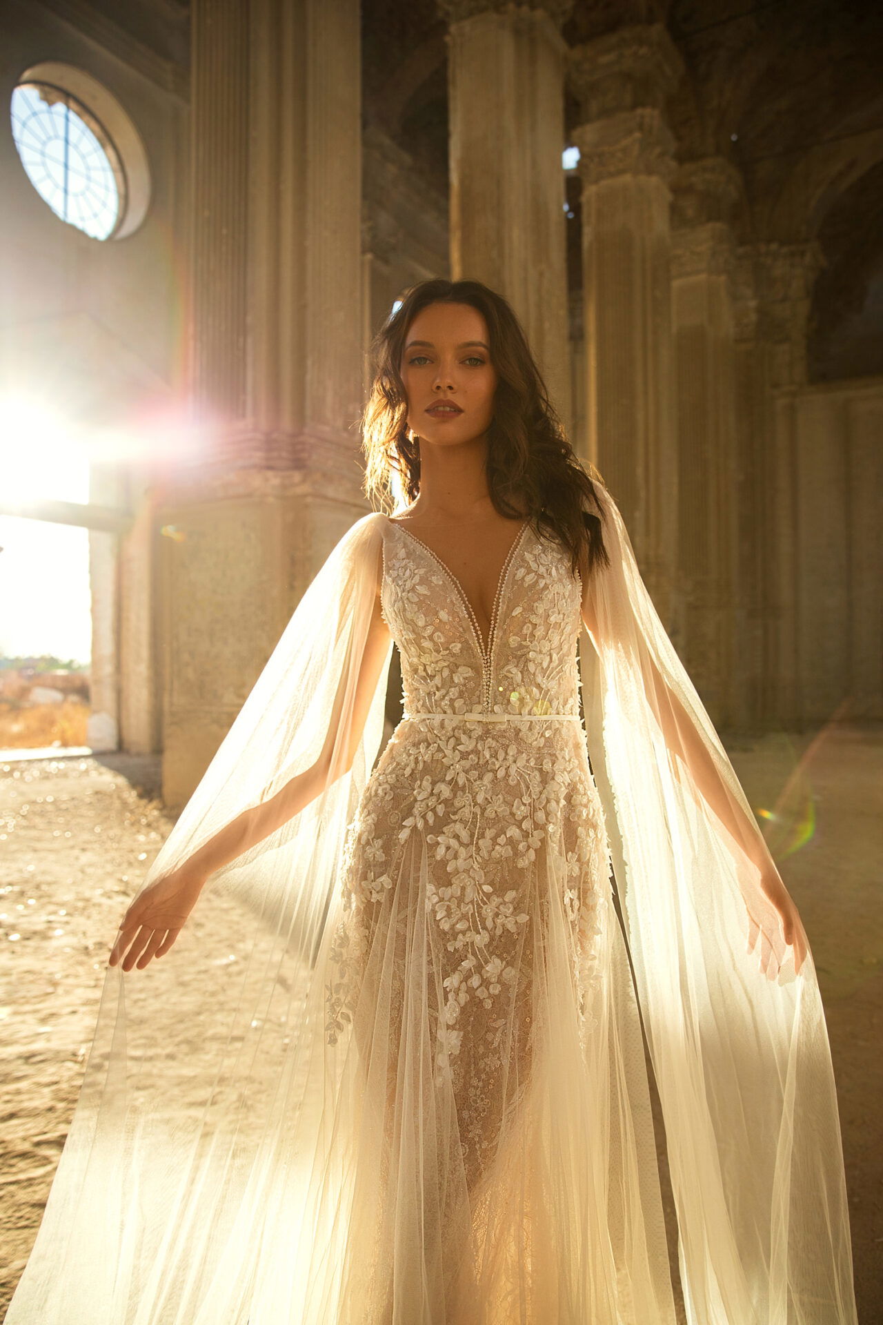 Eine Braut in einem verzierten Kleid mit fließenden Cape-Ärmeln, eingefangen in einem antiken, sonnendurchfluteten Raum mit romantischer Atmosphäre.