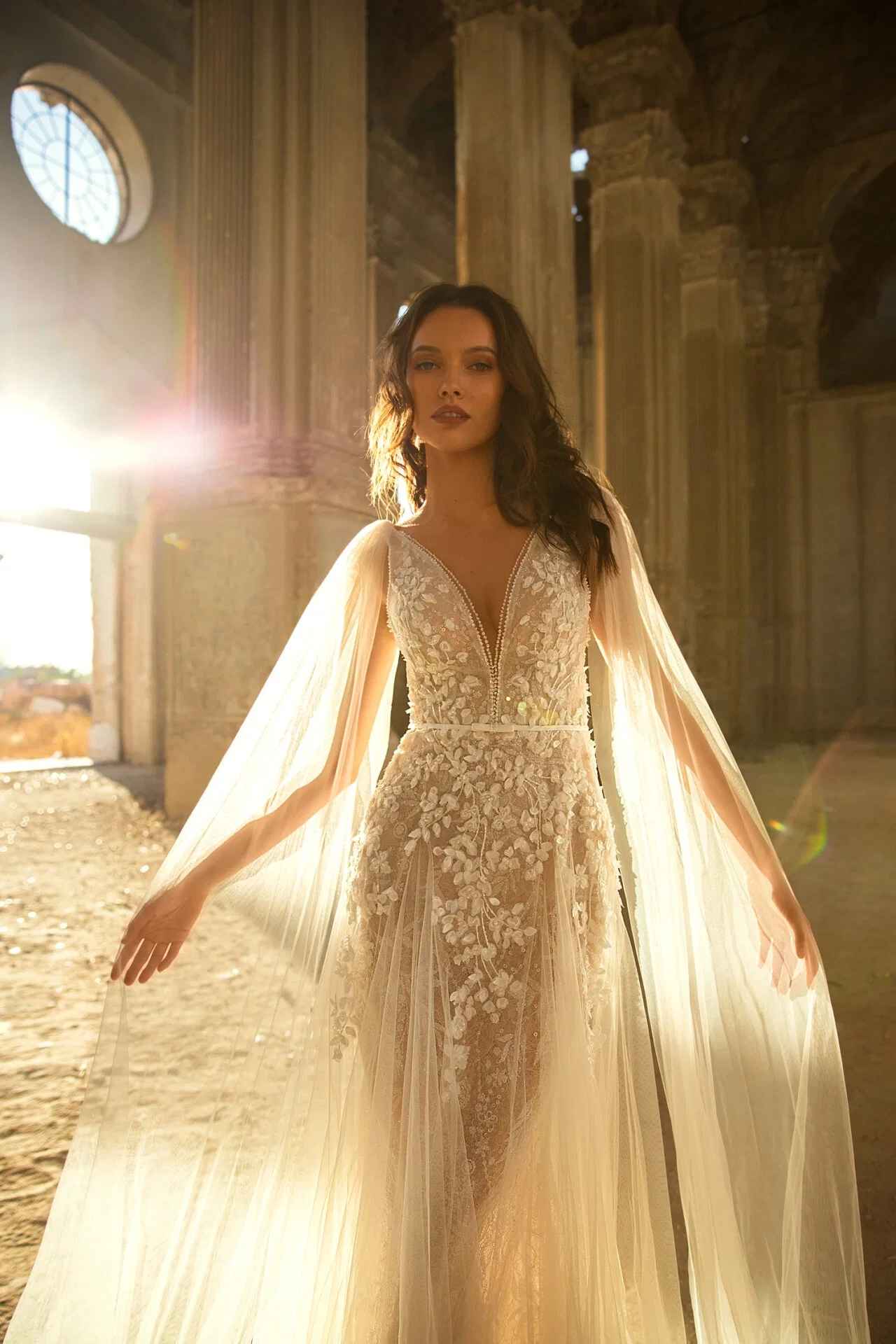 Eine Braut in einem verzierten Kleid mit fließenden Cape-Ärmeln, eingefangen in einem antiken, sonnendurchfluteten Raum mit romantischer Atmosphäre.