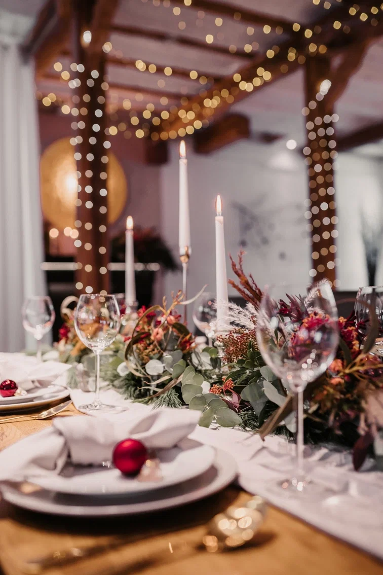 Ein festlicher Tisch mit weihnachtlicher Dekoration, Kerzen und Blumen. Die Lichterketten im Hintergrund sorgen für ein gemütliches Ambiente.