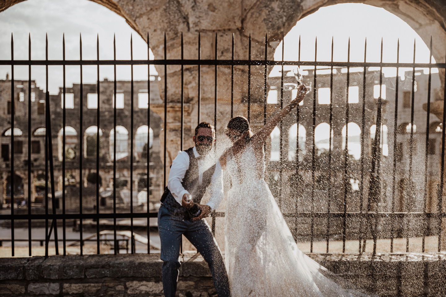 Ein fröhliches Brautpaar feiert, indem es eine Champagnerflasche spritzen lässt. Die Braut in ihrem Hochzeitskleid hebt triumphierend die Arme, während der Bräutigam in Weste und Sonnenbrille lacht.