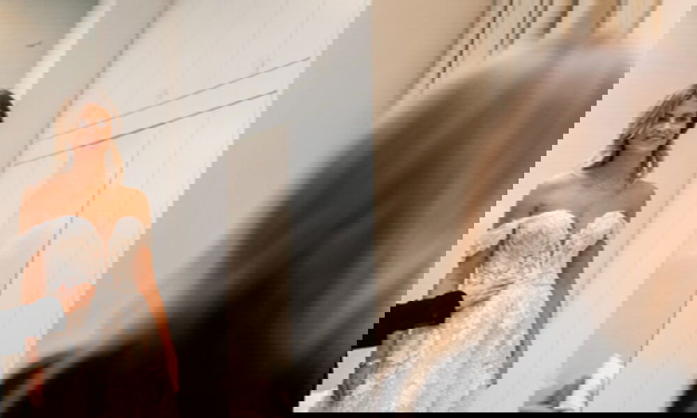 Eine Braut steht in einem wunderschönen, trägerlosen Hochzeitskleid vor einem Spiegel, während eine Stylistin die letzten Anpassungen am Kleid vornimmt. Der helle, modern eingerichtete Raum betont die klassische Eleganz des Brautkleides.