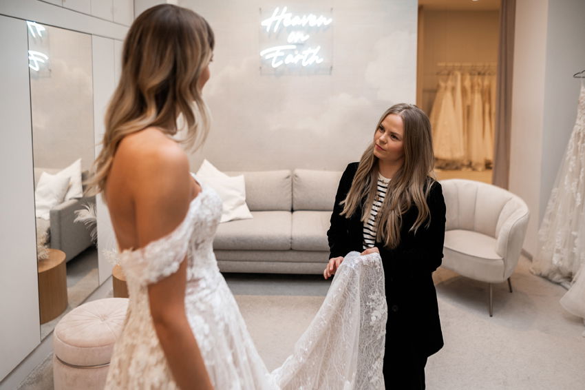 In einem luxuriösen Ankleideraum wird der Saum eines schulterfreien Hochzeitskleides von einer Stylistin gerichtet, während die Braut vor einem Spiegel steht. Die Inneneinrichtung ist hell und modern, mit einem gemütlichen Sofa und einer eleganten Wanddekoration.