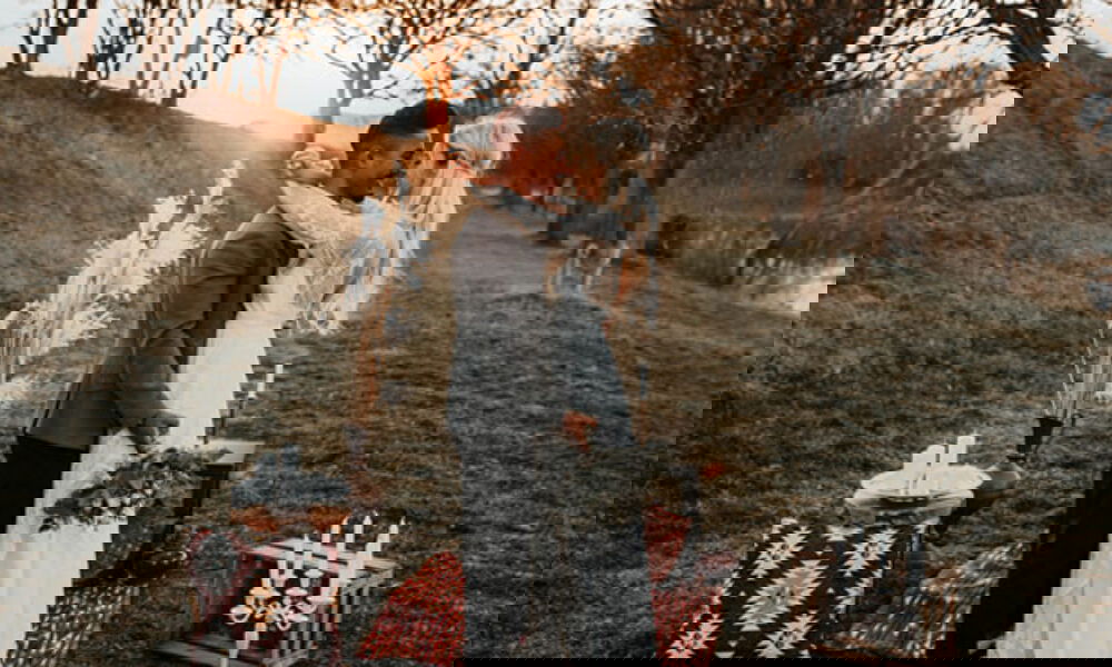 Braut und Bräutigam in inniger Umarmung auf einer Wiese bei Sonnenuntergang. Die Braut in einem weissen Kleid mit langen Spitzenärmeln hält einen kleinen Blumenstrauss, während der Bräutigam im grauen Sakko sie sanft umarmt. Das Paar steht auf einem Teppich in warmen Farben, umgeben von dekorativen Elementen wie einem hohen Trockenblumenarrangement und Kerzen. Die goldene Abendsonne schafft eine stimmungsvolle, verträumte Atmosphäre.