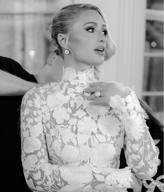 Paris Hilton trägt ein hochgeschlossenes Brautkleid mit filigraner Spitze und floralen Mustern, während sie sich für ihre Hochzeit fertig macht.