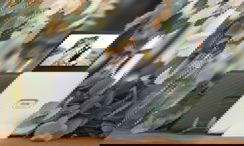 Zwei Instax-Drucker, Schwarz und Weiss, umgeben von Eukalyptus und Trockenblumen auf einem Tisch. Ein Hochzeitsbild eines sich küssenden Paares steht auf dem weissen Drucker und zeigt die liebevolle Stimmung des Moments.