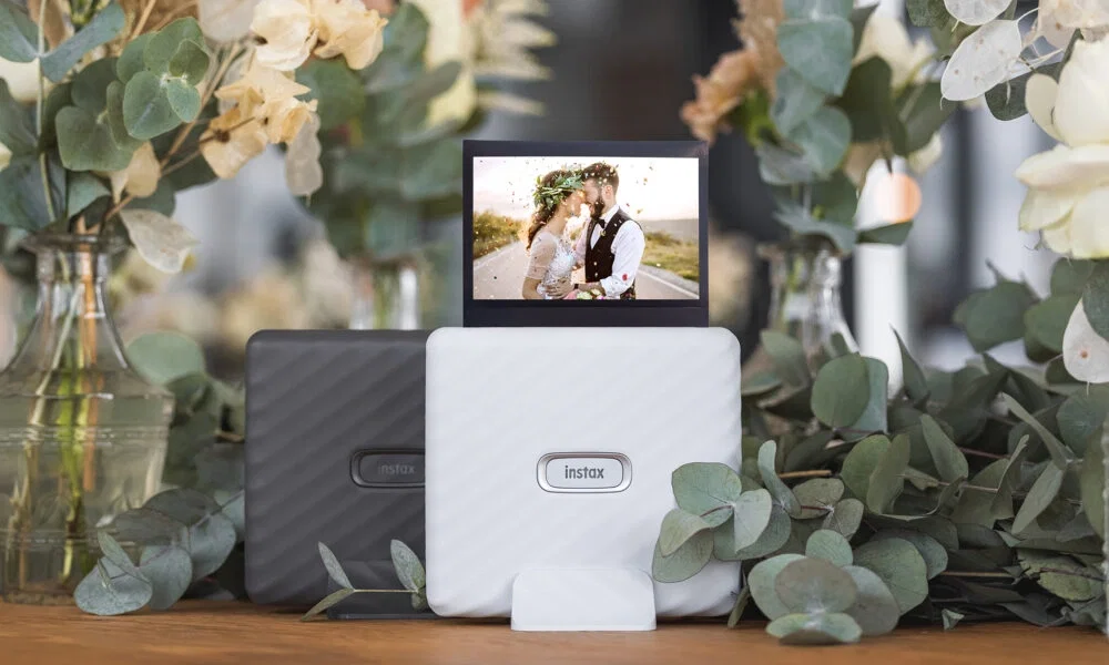 Zwei Instax-Drucker, Schwarz und Weiss, umgeben von Eukalyptus und Trockenblumen auf einem Tisch. Ein Hochzeitsbild eines sich küssenden Paares steht auf dem weissen Drucker und zeigt die liebevolle Stimmung des Moments.