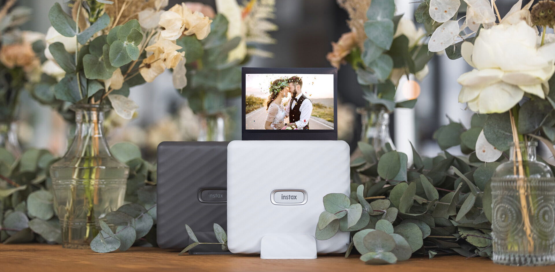 Schwarzer und weisser Instax Link Wide Printer auf einem Tisch mit Eukalyptuszweigen und Blumen im Hintergrund. Auf dem weissen Printer steht ein Hochzeitsbild, das ein Paar beim Kuss zeigt. Die Dekoration mit Trockenblumen und das warme Licht verleihen dem Bild eine romantische Note.
