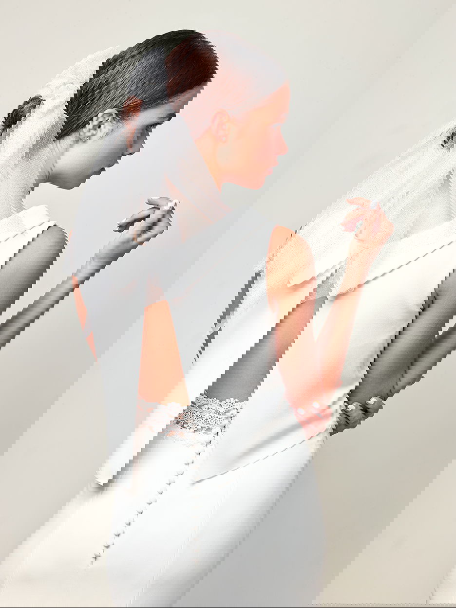 Rückansicht einer Braut, die ein schickes Kleid mit tiefem Rückenausschnitt und Spitzenbändern trägt. Ihr Haar ist in einem Knoten mit Schleier gestylt.