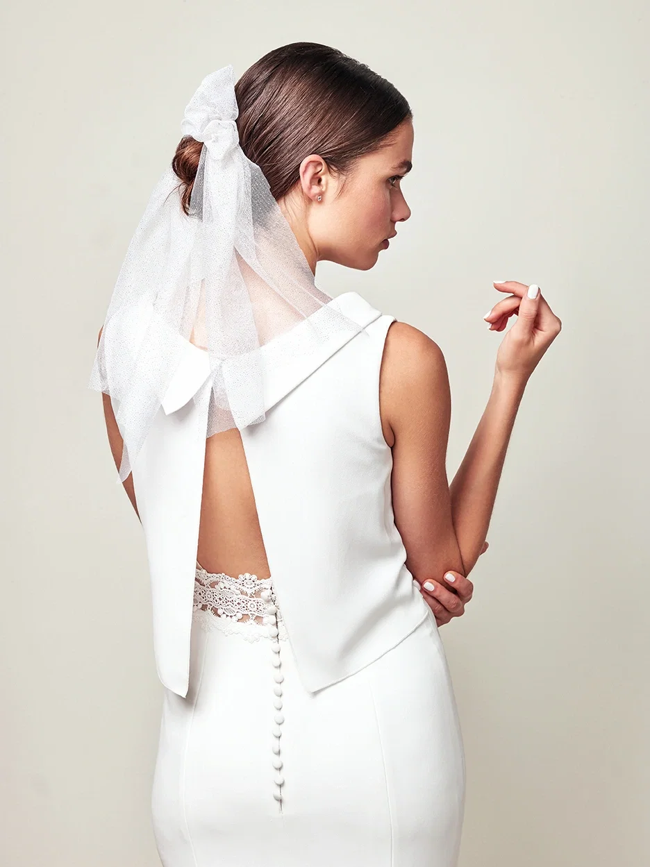 Rückansicht einer Braut, die ein schickes Kleid mit tiefem Rückenausschnitt und Spitzenbändern trägt. Ihr Haar ist in einem Knoten mit Schleier gestylt.