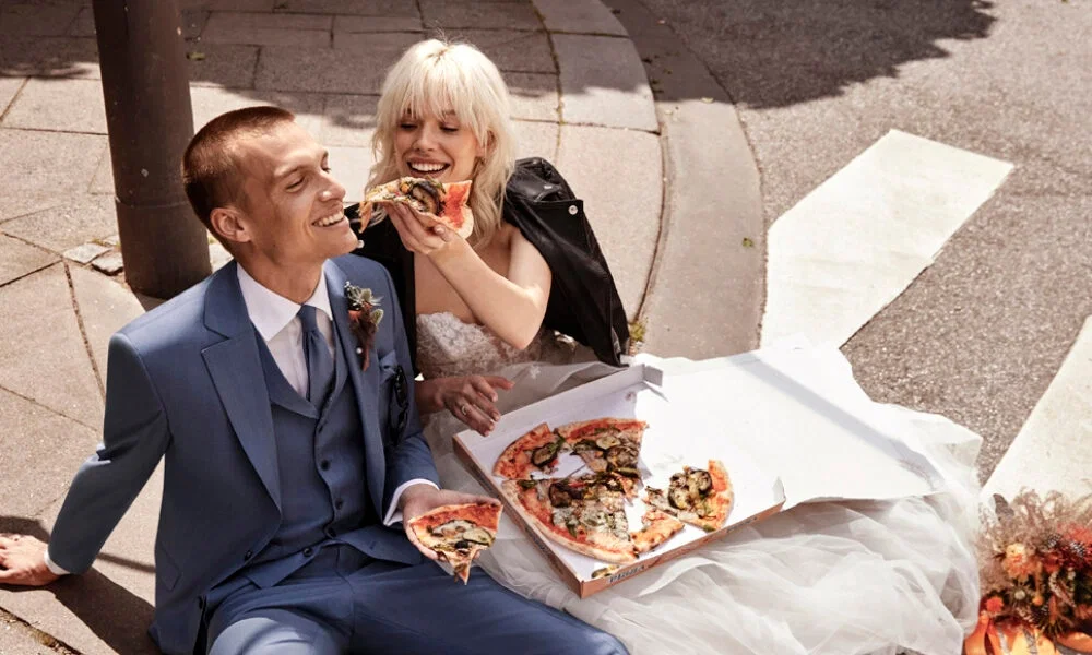 Ein Brautpaar sitzt auf dem Boden und genießt eine Pizza. Die Braut trägt eine schwarze Lederjacke über ihrem Hochzeitskleid, während der Bräutigam einen blauen Anzug trägt.