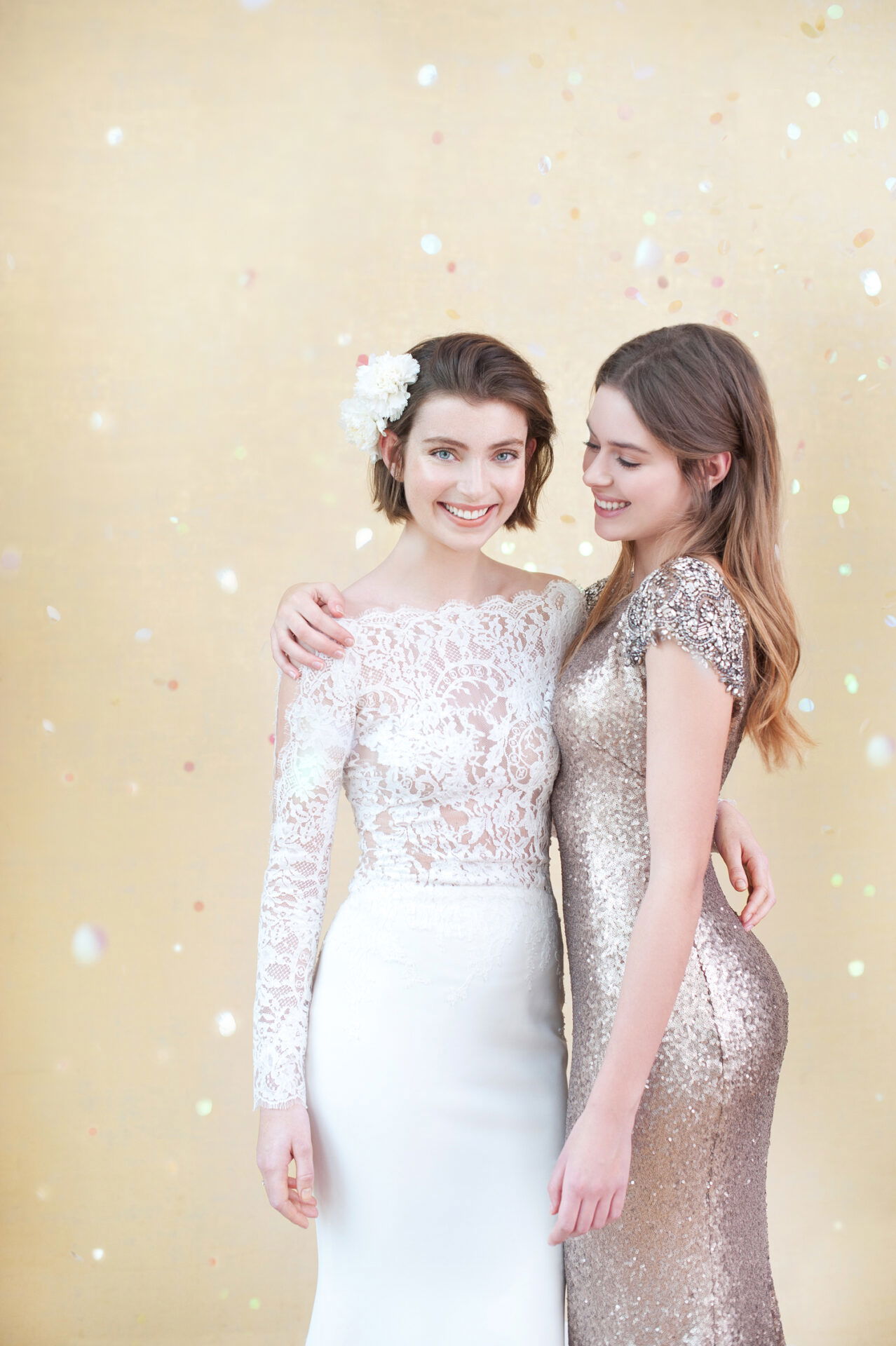 Eine Braut und eine Brautjungfer stehen nebeneinander und lächeln in die Kamera. Die Braut trägt ein weißes Spitzenkleid, die Brautjungfer ein glitzerndes goldfarbenes Kleid. Der Hintergrund ist mit Konfetti gestaltet.