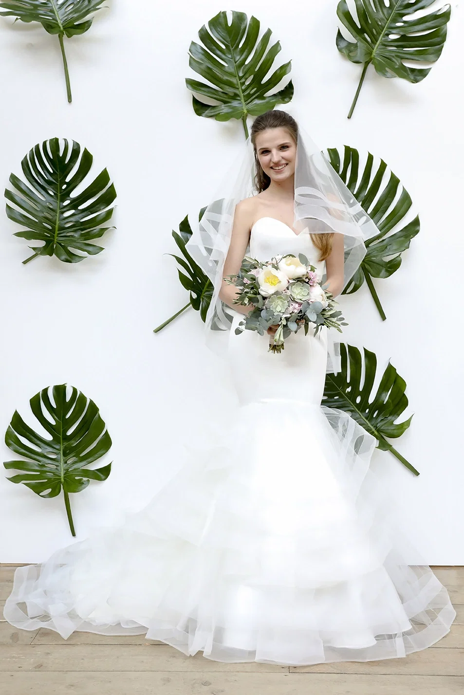 Elegante Braut in einem klassischen, trägerlosen Brautkleid mit ausgestelltem Rock und transparentem Schleier, die einen Blumenstrauß aus weißen und pastellfarbenen Blüten hält. Der Hintergrund besteht aus großen grünen Blättern, die an einer weißen Wand angebracht sind.