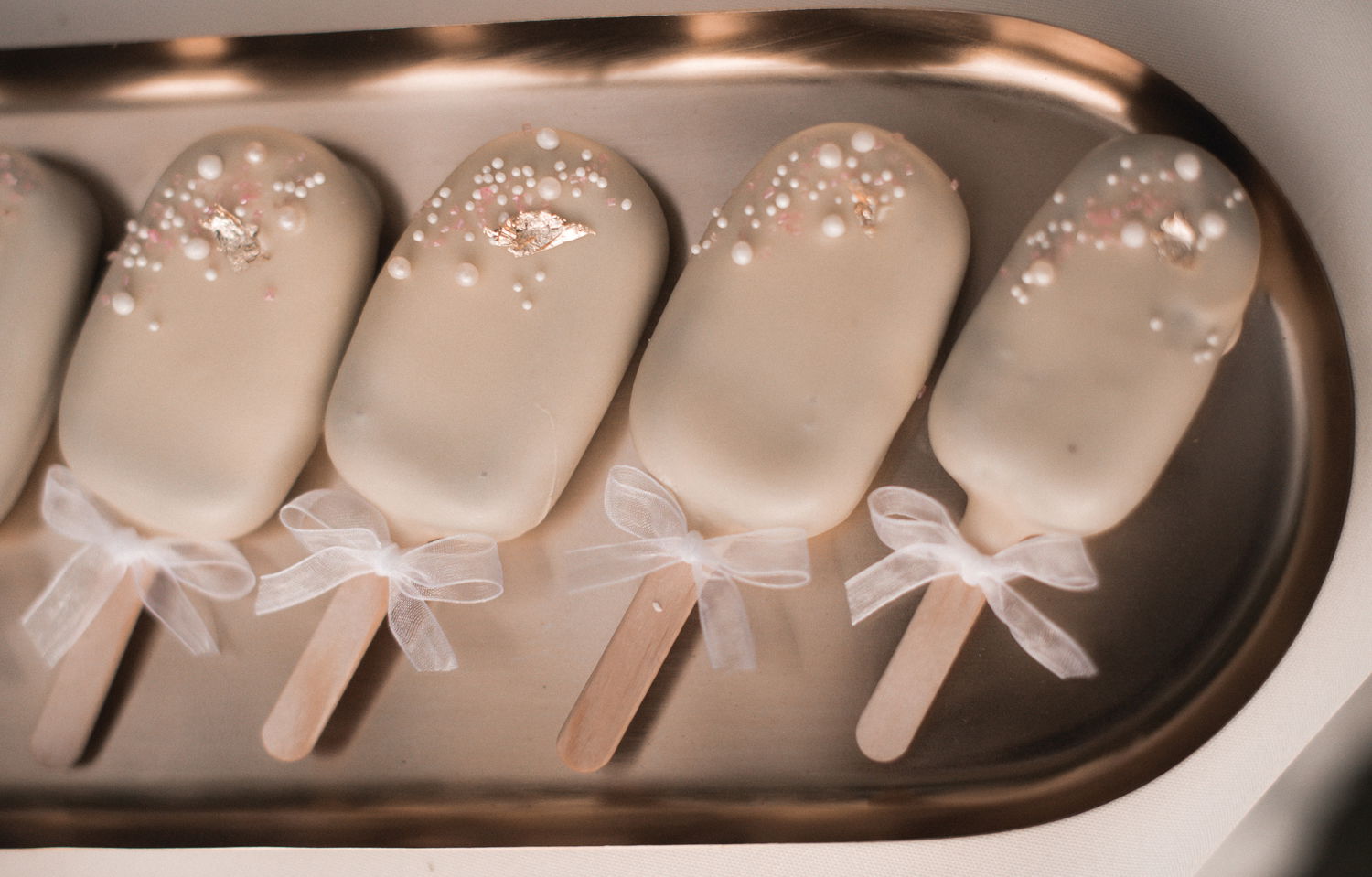 Vier zart dekorierte Cake Popsicles mit glattem, weißem Überzug liegen auf einem goldenen Tablett. Jedes Popsicle ist mit kleinen Perlen und goldenen Flocken verziert, während die Stiele mit weißen Schleifen dekoriert sind, was dem Dessert eine festliche Note verleiht.