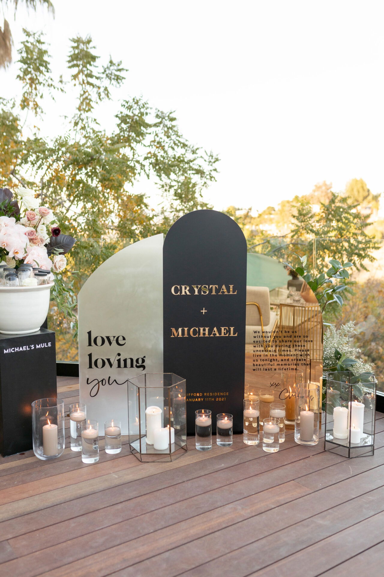 Ein elegantes Hochzeitsschild mit der Aufschrift "Crystal + Michael" und mehreren Kerzen sowie Blumenarrangements, die eine festliche Atmosphäre schaffen.