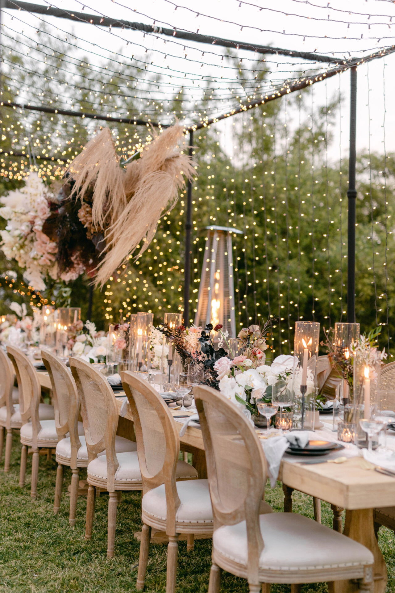 Ein festlich dekorierter Hochzeitstisch im Freien, mit Kerzen und Blumenarrangements, beleuchtet von Lichterketten, die eine gemütliche Atmosphäre schaffen.