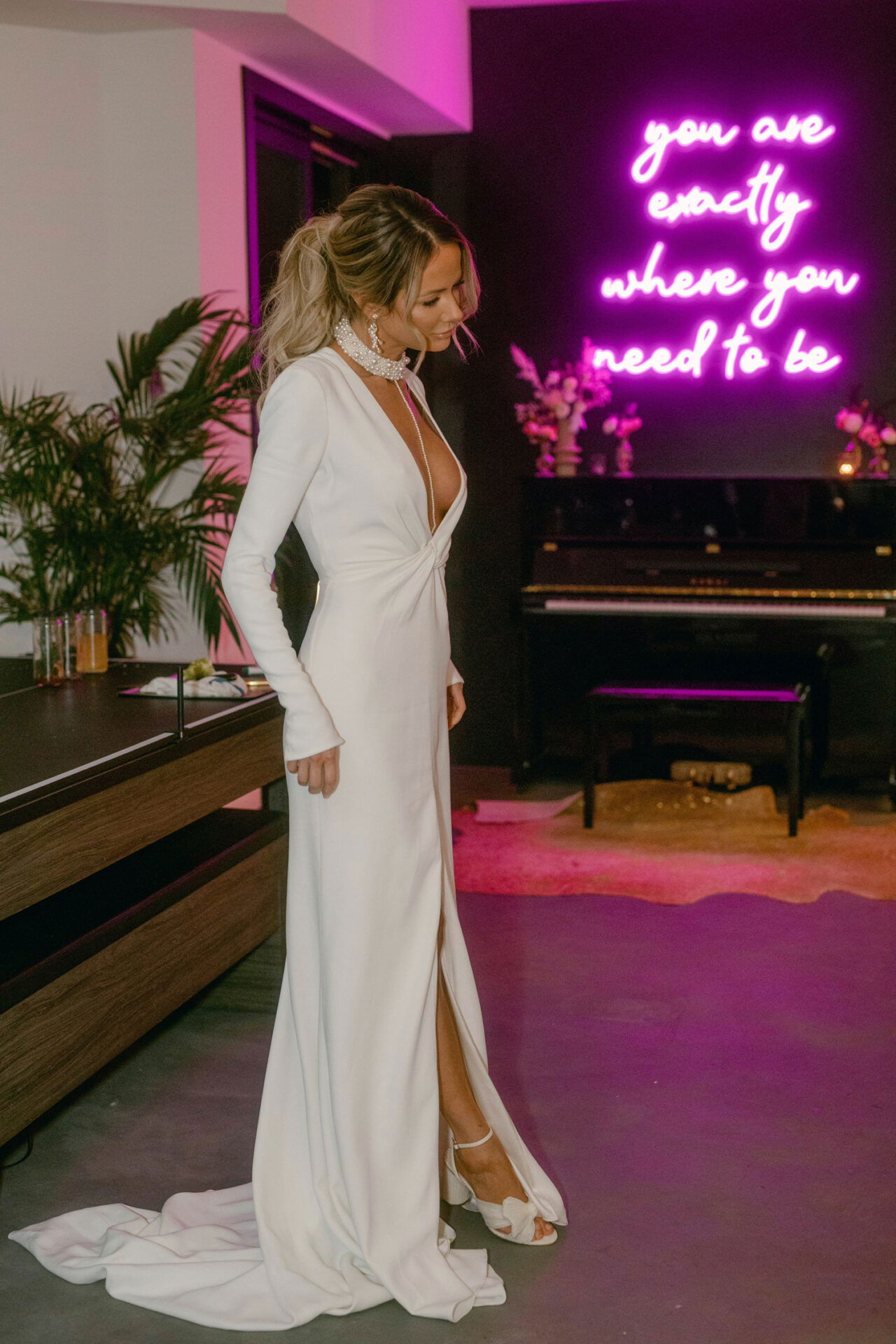 Die Braut in einem langen, tief ausgeschnittenen Hochzeitskleid posiert in einem modernen Raum, beleuchtet von einer pinken Neonwand mit inspirierendem Zitat.