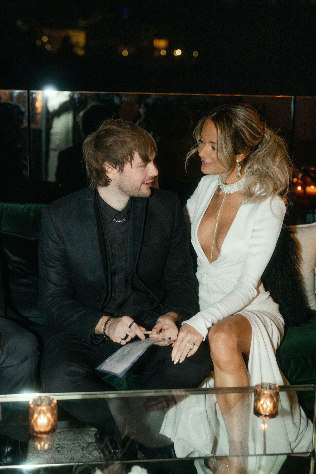 Braut und Bräutigam sitzen auf einem Sofa und genießen einen ruhigen Moment nach der Zeremonie. Sie trägt ein glamouröses, ausgeschnittenes Hochzeitskleid, er einen schlichten schwarzen Anzug.