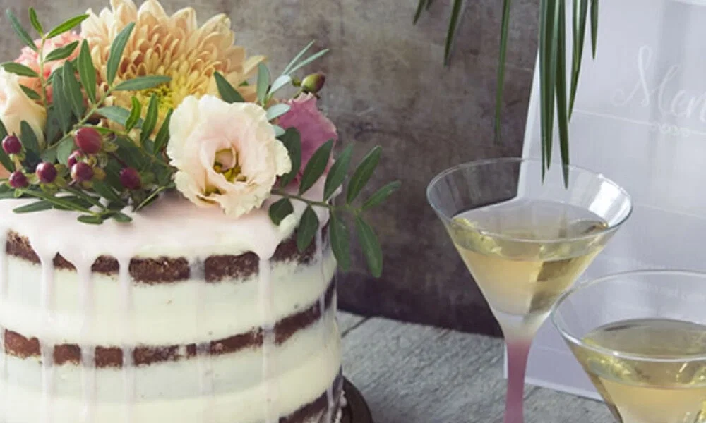 Rustikale Hochzeitstorte mit schlichter, weißer Glasur und tropfenden Details. Sie ist mit Blumen dekoriert und wird von zwei Martini-Gläsern auf einem Tisch ergänzt.