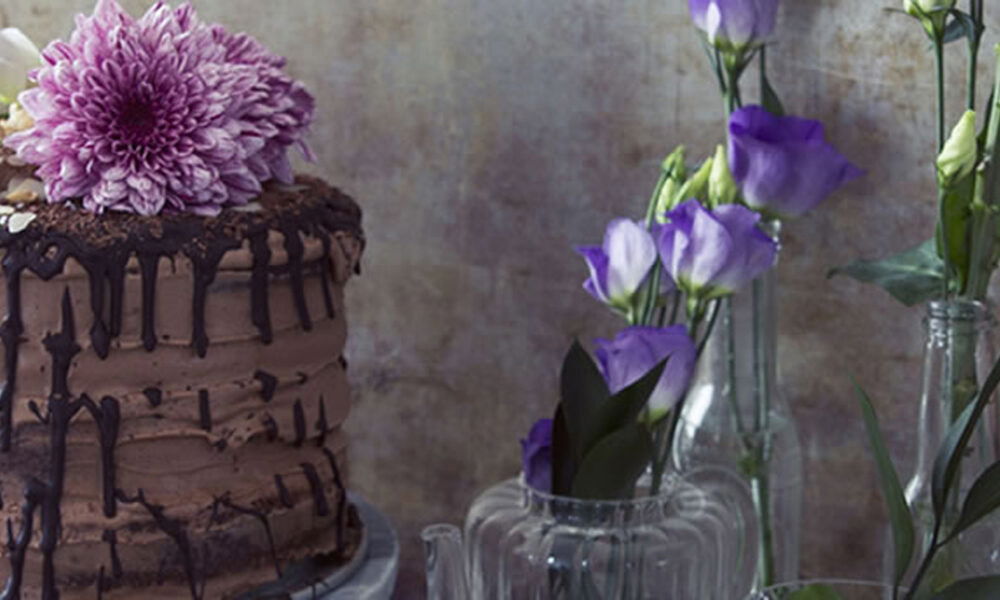 Schichttorte mit Schokoladenganache und Drip-Glasur, geschmückt mit einer violetten Blüte, arrangiert neben stilvollen Glasvasen mit Blumen.