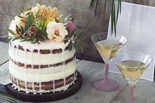 Ein rustikaler Naked Cake, geschichtet mit weißer Glasur und verziert mit Blumen, steht auf einem Tisch. Zwei Martini-Gläser mit einem goldenen Drink ergänzen das festliche Arrangement.