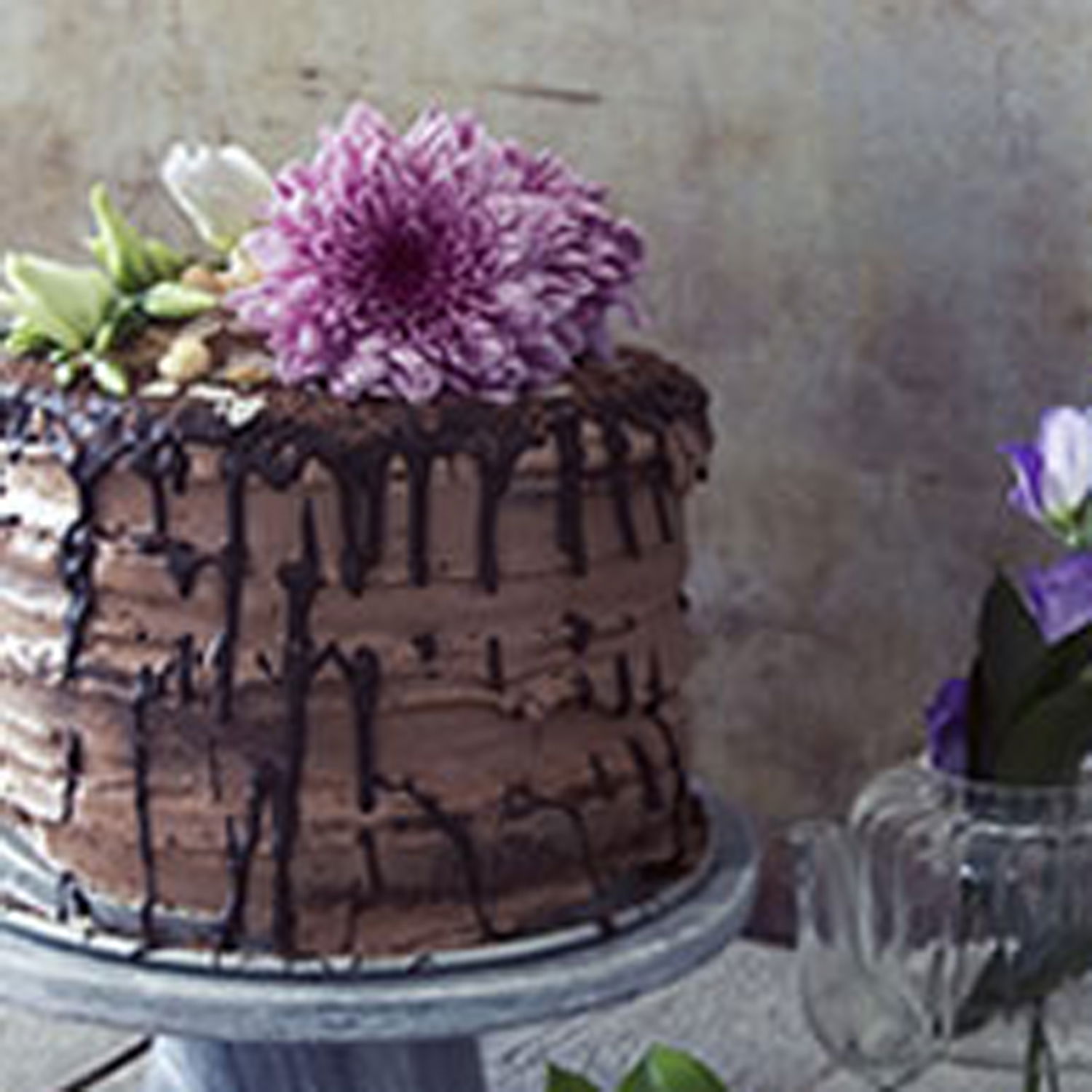 Rustikale Schokotorte mit tropfender Glasur und lila Blütendekor, kunstvoll arrangiert neben filigranen Glasgefäßen mit violetten Blumen.