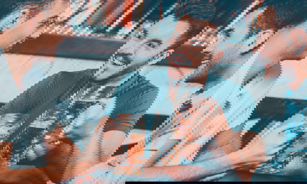 Drei junge Männer sitzen gemeinsam an einem Tisch in einer Bar und prosten sich mit Bierkrügen zu. Sie unterhalten sich gut gelaunt, während hinter ihnen eine stilvolle Theke mit Gläsern und Flaschen zu sehen ist.