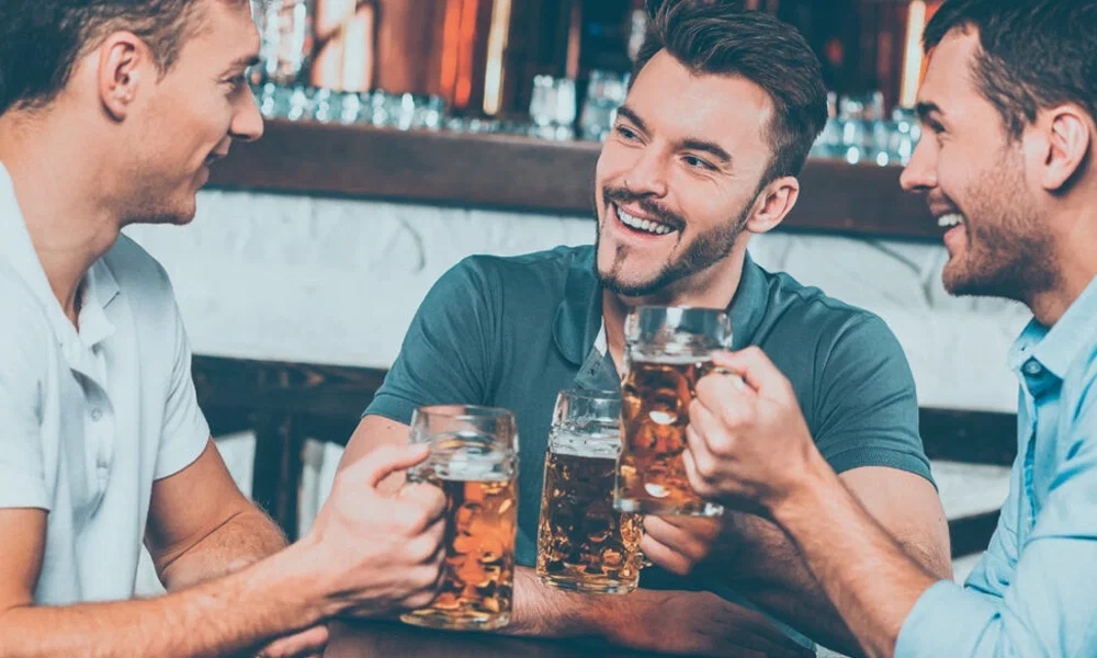Drei junge Männer sitzen gemeinsam an einem Tisch in einer Bar und prosten sich mit Bierkrügen zu. Sie unterhalten sich gut gelaunt, während hinter ihnen eine stilvolle Theke mit Gläsern und Flaschen zu sehen ist.