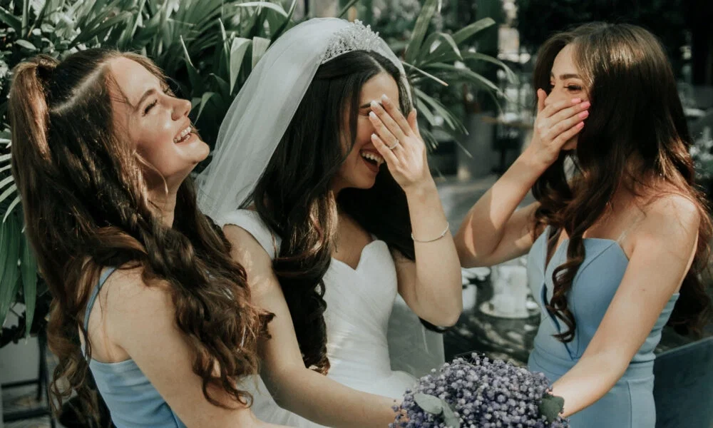 Drei Frauen, die Braut in der Mitte mit einem Schleier und zwei Brautjungfern in blauen Kleidern, lachen ausgelassen. Sie halten sich die Hände lachend vors Gesicht, während sie in einer tropischen Umgebung im Freien stehen.