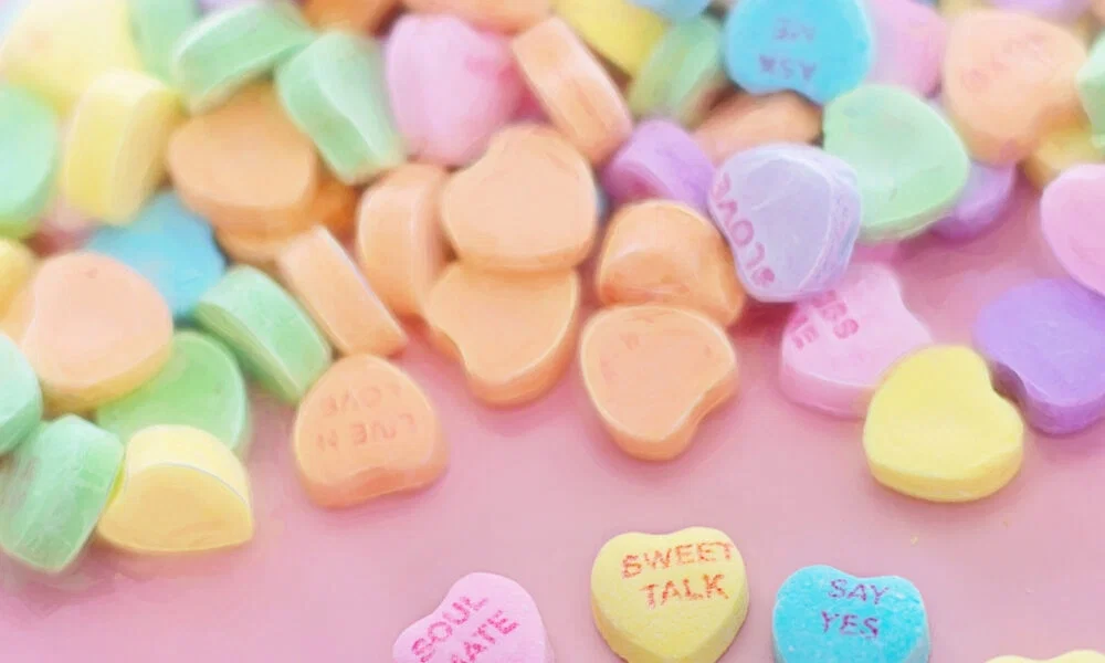 Pastellfarbene Herzbonbons mit liebevollen Aufschriften wie „Sweet Talk“ und „Love“ liegen auf einem rosa Untergrund und verbreiten einen süßen, verspielten Charme.