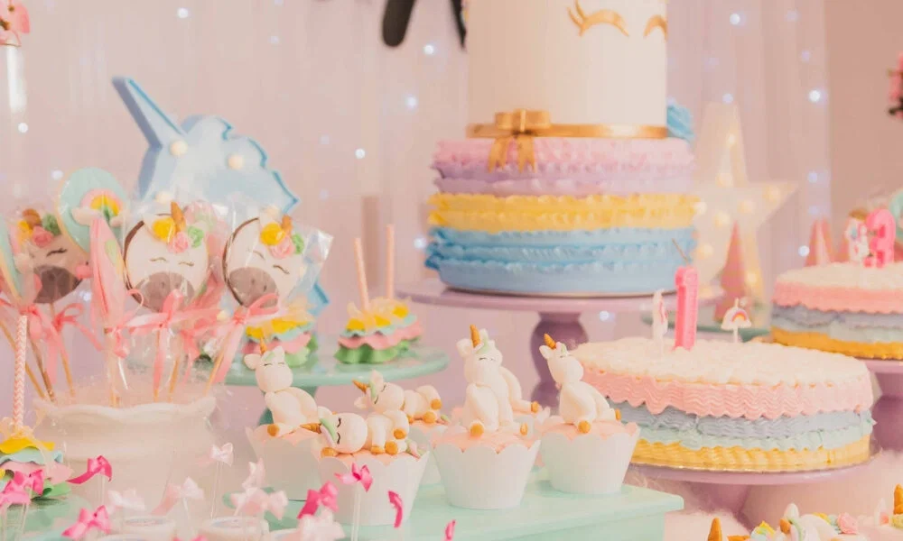 Ein Desserttisch im Einhorn-Stil mit bunten Torten, Cupcakes und Einhorn-Lollies. Pastellfarben und niedliche Einhornfiguren dominieren die liebevolle Dekoration.