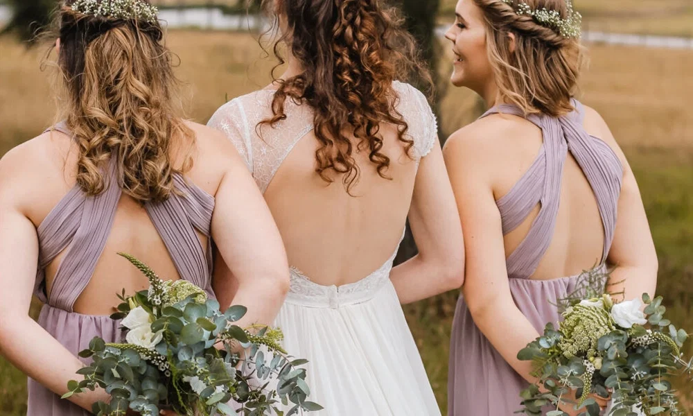 Die Rückansicht einer Braut und zwei Brautjungfern in einer ländlichen Umgebung. Die Braut trägt ein elegantes Spitzenkleid, während die Brautjungfern fliederfarbene Kleider tragen. Alle halten Blumensträuße und tragen florale Haarkränze.