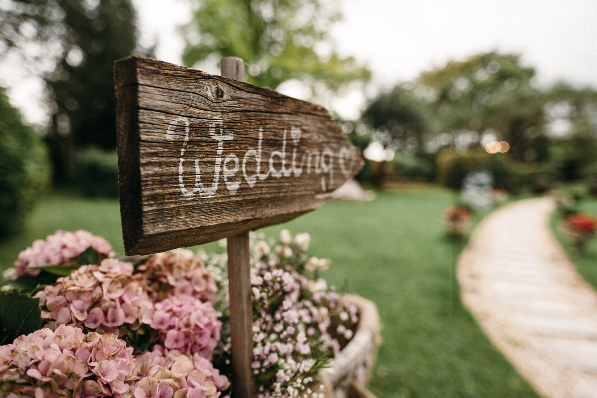 Ein handbeschrifteter Holzpfeil mit 'Wedding' weist den Weg zur Hochzeitslocation, während üppige rosa Hortensien im Vordergrund blühen.