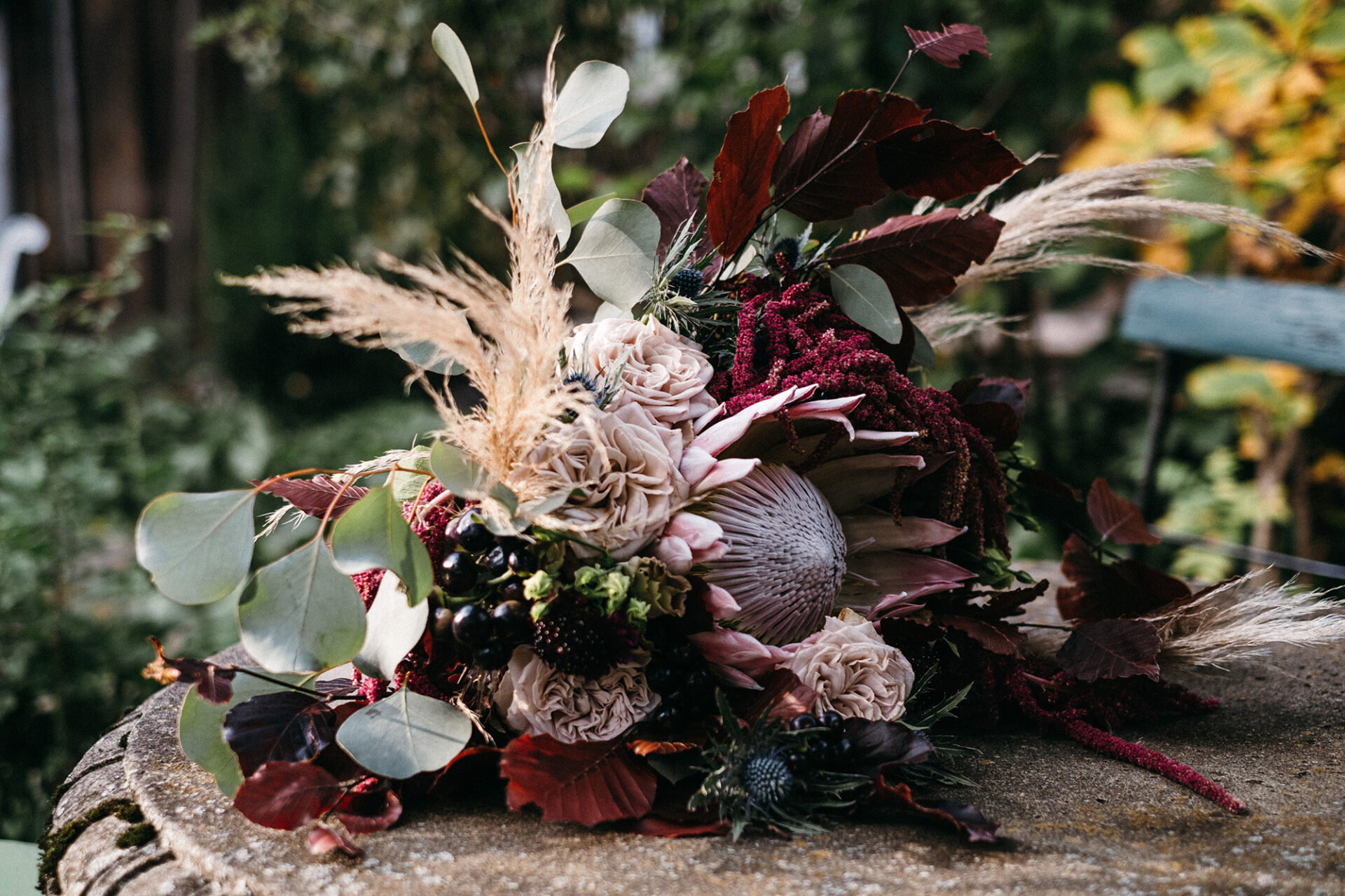 Rustikales Blumengesteck mit Protea, Brombeeren und Eukalyptus, das durch Pampasgras und dunkle Akzente ergänzt wird, arrangiert auf einem Holzuntergrund.