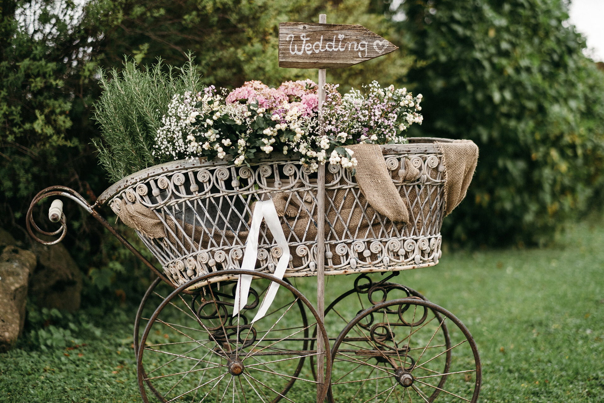 Vintage-inspirierter Kinderwagen, dekoriert mit frischen Blumen und Jutestoff, zeigt den Hochzeitsweg in einem grünen Garten und verleiht nostalgischen Charme.