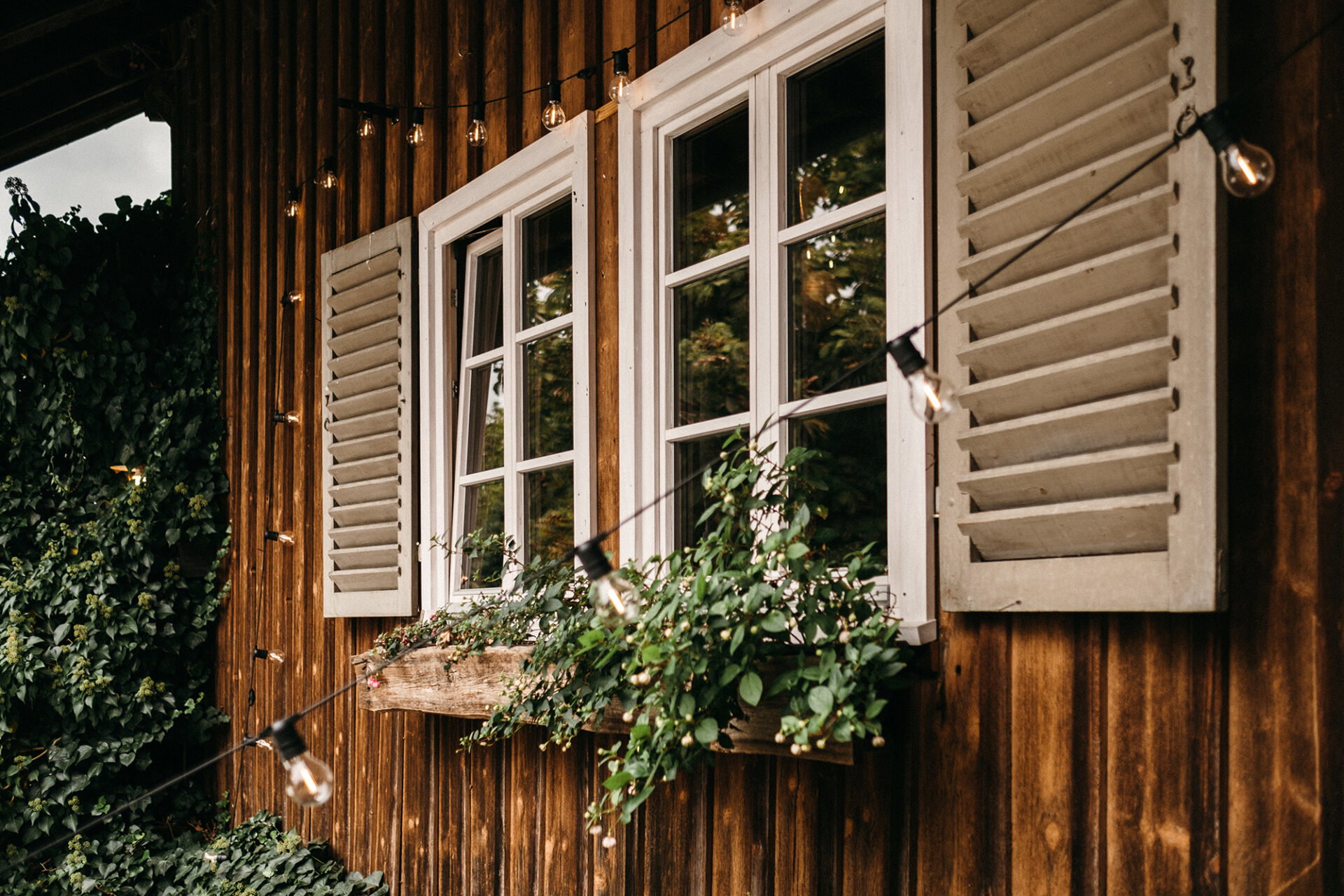 Romantisches Landhaus mit weißen Fensterrahmen und hölzernen Fensterläden, geschmückt mit einer Lichterkette und Blumen, ideal für eine Hochzeit im Freien.