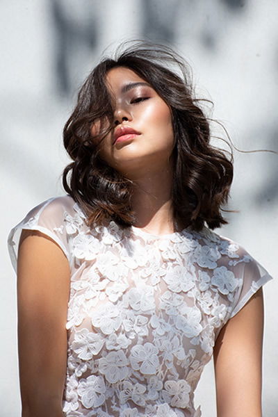 Junge Frau mit schulterlangem, welligem Haar trägt ein florales Spitzenkleid. Mit geschlossenen Augen und im Sonnenlicht wirkt sie in einem Moment purer Entspannung.