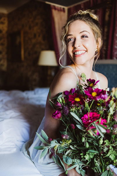 Fröhliche Braut mit einem Blumenstrauß in kräftigen Violetttönen, die auf einem Bett sitzt. Ihr strahlendes Lächeln und die Umgebung erzeugen eine intime Atmosphäre.